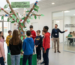 Un atrio scolastico dove un gruppo di studenti osserva un'installazione creativa a forma di albero chiamata "L'Ecosistema dell'Apprendimento". Sullo sfondo, un'aula vetrata con l'insegna "Laboratorio L2 - Spazio di Parole" mostra una lezione in piccoli gruppi, illustrando il modello didattico pull-out per l'italiano L2.